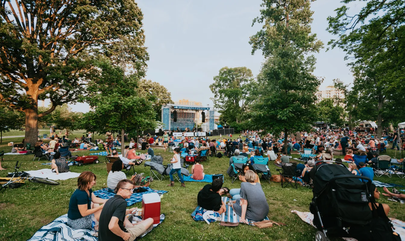 A large crowd gathered on blankets and lawn chairs in a park, watching an outdoor concert on a stage surrounded by trees.