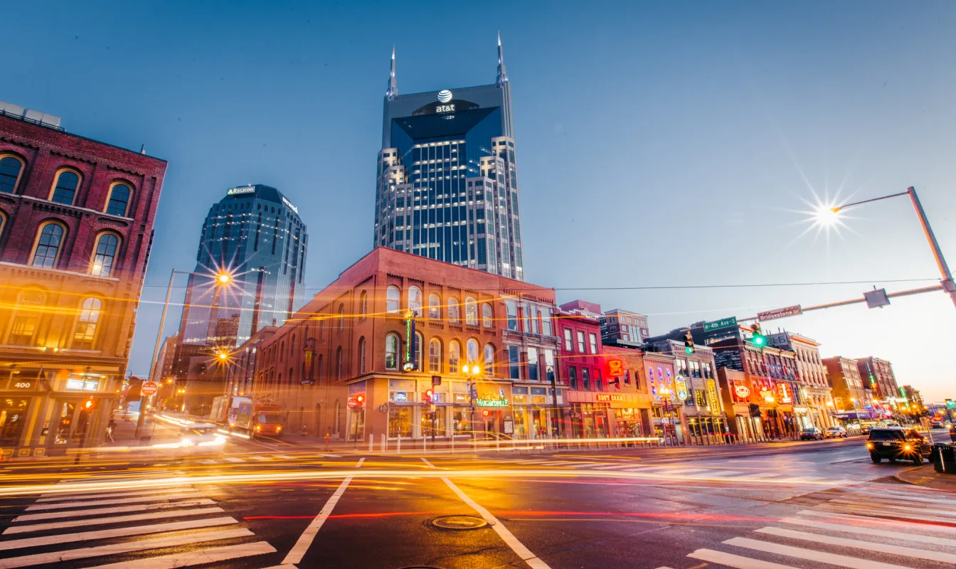 Downtown Nashville street at dusk with neon-lit buildings and the AT&T Tower in the background.