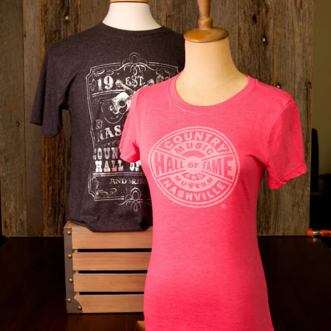 Two mannequins displaying Country Music Hall of Fame T-shirts — pink fitted tee with circular logo in front, dark gray graphic tee behind.