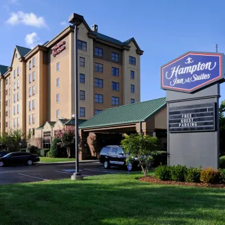 Exterior of Hampton Inn & Suites with green roof and roadside sign reading “Hampton Inn & Suites” and “Free Guest Parking”.