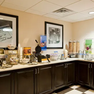 Hotel breakfast station with waffle maker, cereal dispensers, juice machine, and hot items on black cabinetry.