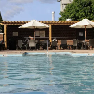 Outdoor hotel pool facing a brick pergola with shaded seating and white umbrellas.
