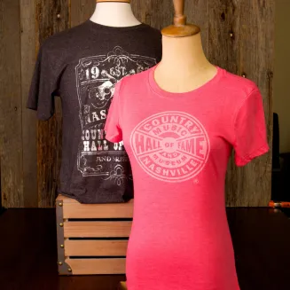 Two mannequins displaying Country Music Hall of Fame T-shirts — pink fitted tee with circular logo in front, dark gray graphic tee behind.