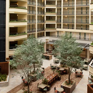 Hotel atrium dining area with tall indoor trees, tables and chairs, and multi-story balconies.