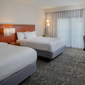 Hotel guest room with two queen beds, desk, and window with sheer curtains.