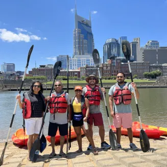 A group of kayakers pose for a photo on the bank of the Cumberland River with the Nashville skyline in the background. Nashville, TN.