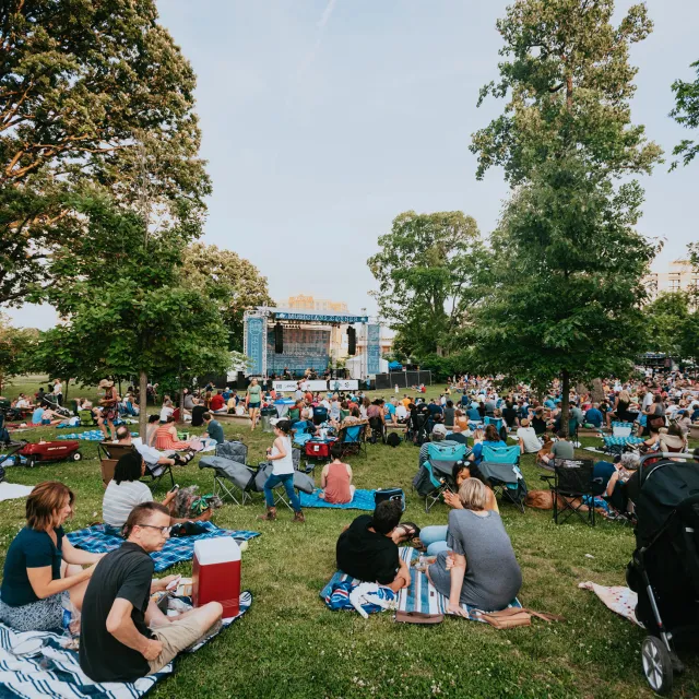 A large crowd gathered on blankets and lawn chairs in a park, watching an outdoor concert on a stage surrounded by trees.