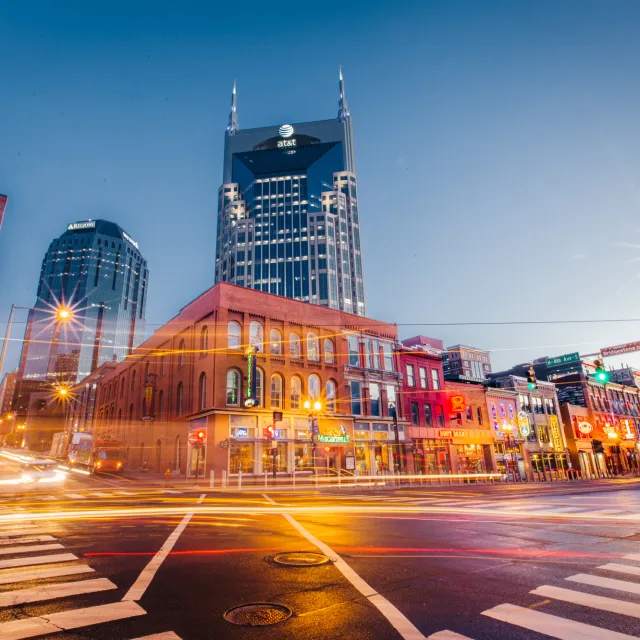 Downtown Nashville street at dusk with neon-lit buildings and the AT&T Tower in the background.